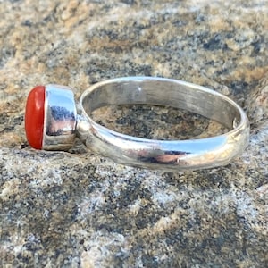 Pu&ograve; includere: Un anello d'argento con una pietra preziosa di corallo rosso. L'anello &egrave; posto su una roccia grigia e ruvida.