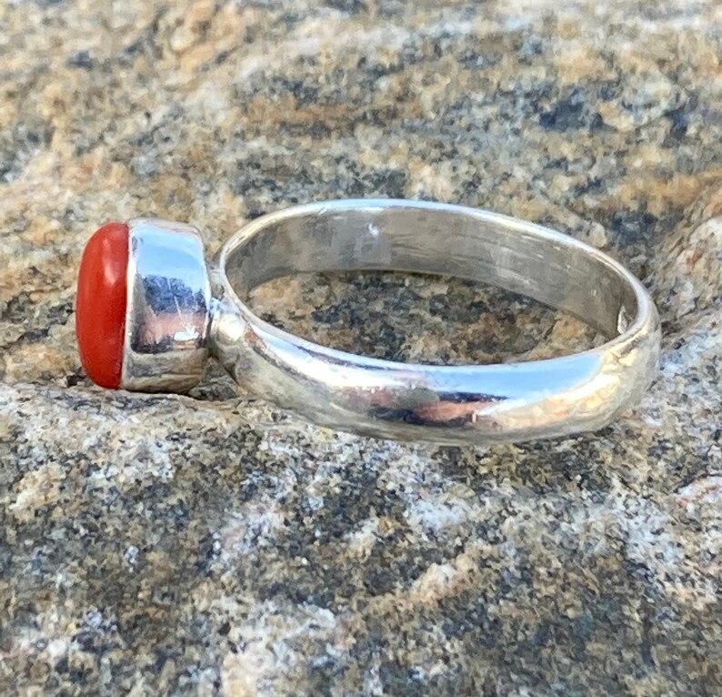 Pu&ograve; includere: Un anello d'argento con una pietra preziosa di corallo rosso. L'anello &egrave; posto su una roccia grigia e ruvida.