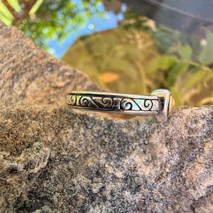 May include: A silver-toned metal ring featuring a continuous band with an embossed, dark-oxidized scrollwork pattern. The ring has a smooth, polished finish along the edges and a small, distinct gold-toned accent piece at one end of the band.