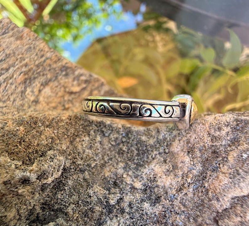 May include: A silver-toned metal ring featuring a continuous band with an embossed, dark-oxidized scrollwork pattern. The ring has a smooth, polished finish along the edges and a small, distinct gold-toned accent piece at one end of the band.