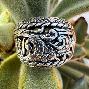 May include: A silver ring with an intricate, ornate design. The ring is made of a filigree pattern with a textured surface.