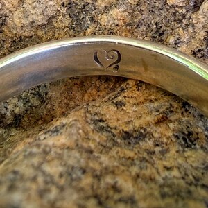 May include: A polished silver-toned metal ring band featuring a smooth, reflective surface. The interior of the band is engraved with a small, stylized heart symbol that includes a delicate swirl detail at the top and a single dot at the base.