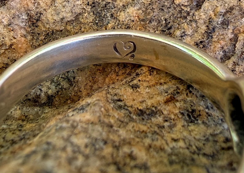May include: A polished silver-toned metal ring band featuring a smooth, reflective surface. The interior of the band is engraved with a small, stylized heart symbol that includes a delicate swirl detail at the top and a single dot at the base.