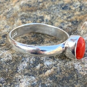 Pu&ograve; includere: Un anello d'argento con una pietra ovale di colore rosso brillante. L'anello &egrave; posto su una superficie ruvida e testurizzata.