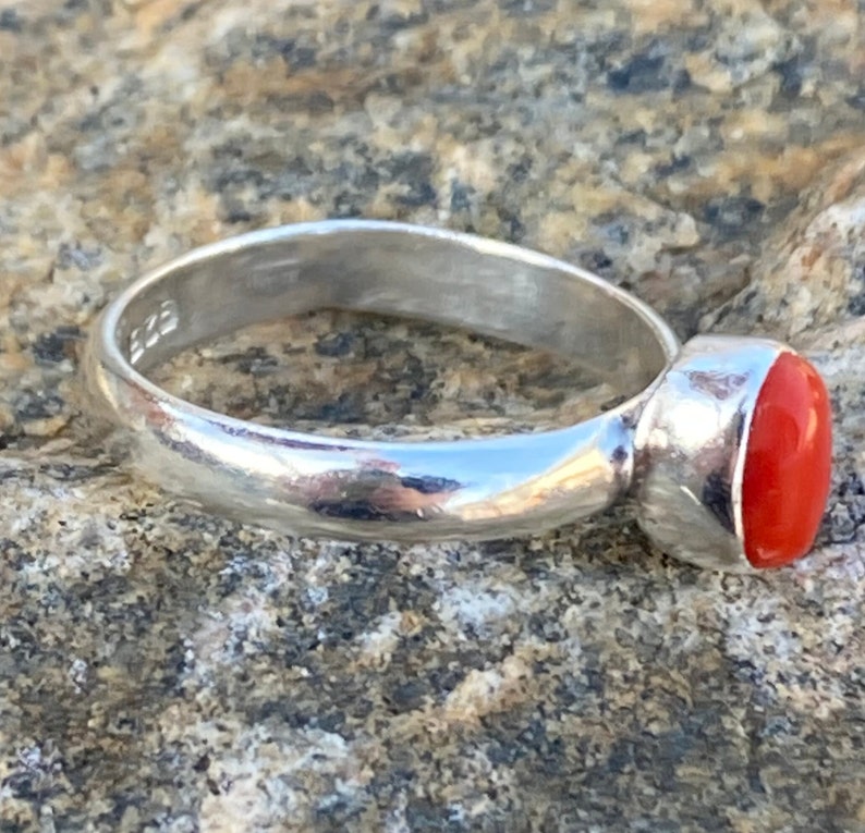 Pu&ograve; includere: Un anello d'argento con una pietra ovale di colore rosso brillante. L'anello &egrave; posto su una superficie ruvida e testurizzata.