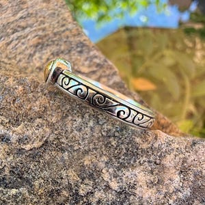 May include: A silver ring featuring a continuous band with an embossed scrollwork pattern and a small, rounded gold-toned accent at the top. The metal surface displays a polished finish with darkened recessed areas to highlight the intricate design.