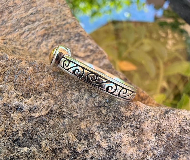 May include: A silver ring featuring a continuous band with an embossed scrollwork pattern and a small, rounded gold-toned accent at the top. The metal surface displays a polished finish with darkened recessed areas to highlight the intricate design.