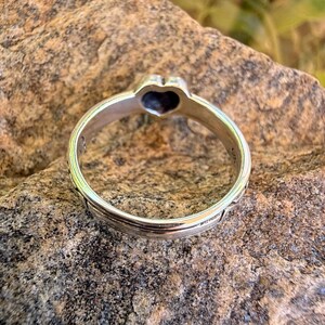 May include: A silver-toned metal ring featuring a heart-shaped setting at the top. The band has a smooth, polished finish with a subtle grooved line running along the center. The interior of the band shows a small hallmark stamp near the heart.