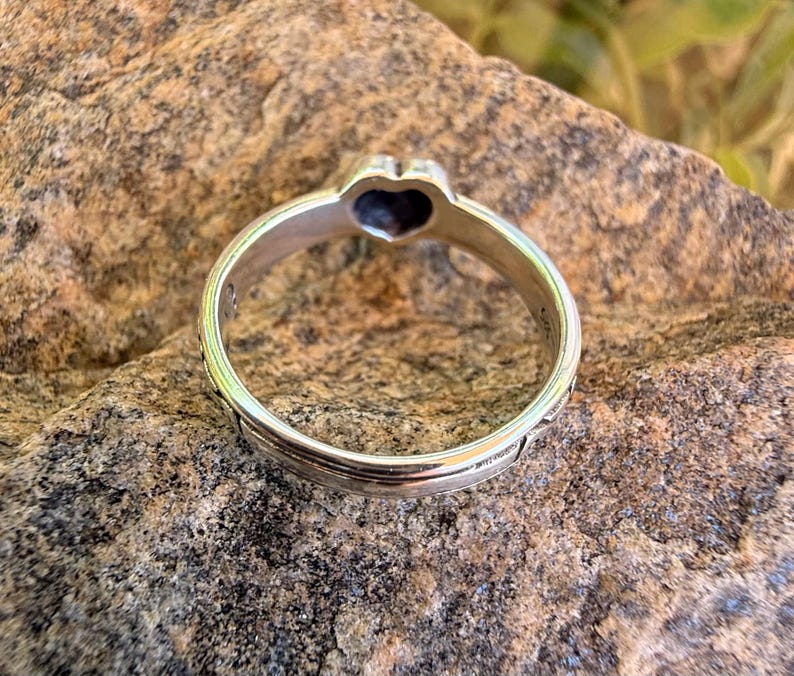 May include: A silver-toned metal ring featuring a heart-shaped setting at the top. The band has a smooth, polished finish with a subtle grooved line running along the center. The interior of the band shows a small hallmark stamp near the heart.