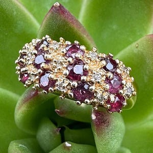 May include: A gold ring with a cluster of deep red gemstones and small diamonds. The ring is set against a backdrop of green succulent leaves, creating a contrast of colors and textures. The ring's design is intricate and eye-catching.