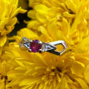 Peut inclure: Une bague en or avec une pierre précieuse rouge foncé, ronde, au centre, flanquée de deux pierres précieuses claires, triangulaires, plus petites. La bague est posée sur un fond de fleurs jaunes vives. Le design de la bague présente des formes géométriques.