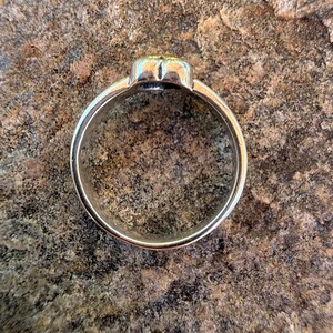 May include: A silver-toned metal ring featuring a smooth, circular band and a raised, rounded top section with a subtle gold-colored accent. The ring is captured from a top-down perspective, showcasing its polished metallic finish and simple design.