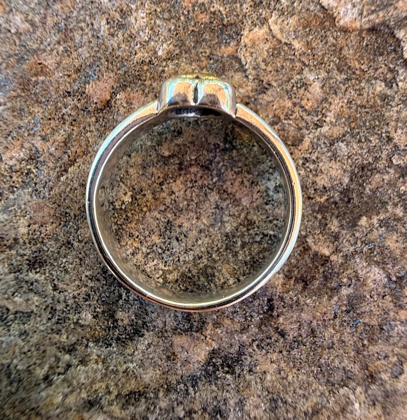 May include: A silver-toned metal ring featuring a smooth, circular band and a raised, rounded top section with a subtle gold-colored accent. The ring is captured from a top-down perspective, showcasing its polished metallic finish and simple design.