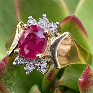 Peut inclure: Une bague en or avec un rubis rouge foncé taille ovale au centre. Le rubis est entouré de deux groupes de petits diamants ronds et transparents sertis sur du métal blanc. L'anneau est en or jaune poli avec un design large et effilé.