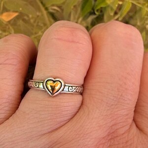 May include: A silver-toned metal ring featuring a central polished gold-toned heart element. The band of the ring displays an embossed scrollwork pattern that wraps around the circumference, providing a textured contrast to the smooth heart.