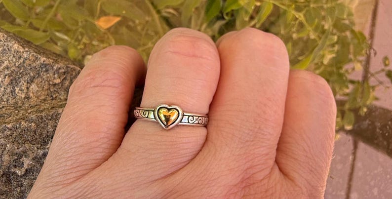 May include: A silver-toned metal ring featuring a central polished gold-toned heart element. The band of the ring displays an embossed scrollwork pattern that wraps around the circumference, providing a textured contrast to the smooth heart.