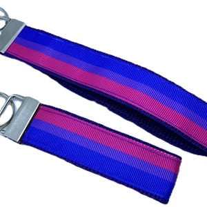 May include: Two keychains with a blue, pink, and blue striped design. The keychains have a silver metal clasp and a key ring. The keychains are a wristlet style, with a loop of fabric for the wrist.