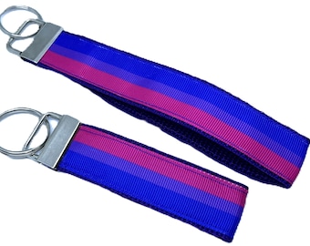 May include: Two keychains with a blue, pink, and blue striped design. The keychains have a silver metal clasp and a key ring. The keychains are a wristlet style, with a loop of fabric for the wrist.