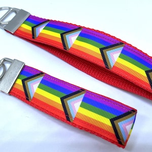 May include: Two red keychains with a rainbow stripe design and silver key rings. The rainbow stripes include the colors of the LGBTQ+ pride flag. The keychains have a metal clasp.