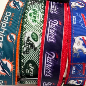 Op de afbeelding: Vier verschillende hondenriemen met teamlogo's. De riemen zijn turquoise met een dolfijn, groen met een Jets-logo, paars met het Patriots-logo en blauw met het Bills-logo.