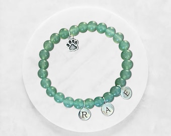 May include: A stretch bracelet made of round, light green translucent beads. The bracelet features a silver-toned metal charm shaped like a paw print and three circular silver-toned metal charms, each engraved with one of the letters R, A, and E.