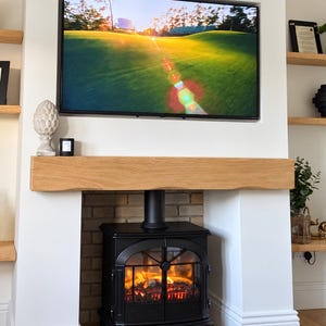 May include: A black cast iron wood-burning stove sits inside a white fireplace alcove with a natural oak mantelpiece. Above the mantel, a flat-screen television displays a golf course landscape. Wooden shelves flank the fireplace with decor.