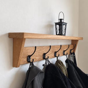 Op de afbeelding: Een aan de muur gemonteerde kapstok van lichtbruin hout met een platte bovenplank en vijf zwarte metalen haken. Verschillende jassen in grijze, zwarte en olijfgroene tinten hangen aan de haken. Een kleine zwarte metalen lantaarn staat op de plank.