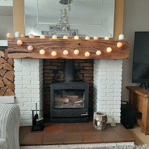 May include: A white brick fireplace with a wood-burning stove and a wooden mantel. The mantel is decorated with a string of white lights. There is a pile of firewood in the background.