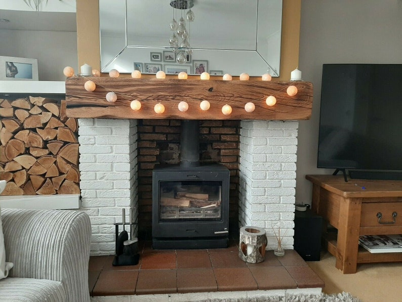 May include: A white brick fireplace with a wood-burning stove and a wooden mantel. The mantel is decorated with a string of white lights. There is a pile of firewood in the background.