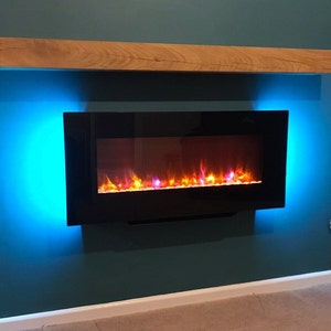 May include: A black electric fireplace with a realistic flame effect is mounted on a teal wall. The fireplace is surrounded by blue LED lights. A wooden mantel sits above the fireplace.
