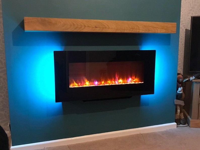 May include: A black electric fireplace with a realistic flame effect is mounted on a teal wall. The fireplace is surrounded by blue LED lights. A wooden mantel sits above the fireplace.