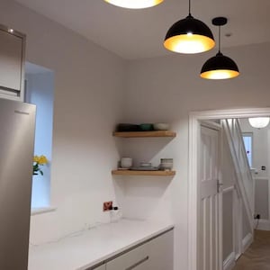May include: Interior kitchen shot with two black pendant lights with gold interiors. The kitchen features white walls, a white countertop, and wooden shelves with dishware. A stainless steel refrigerator is visible on the left.