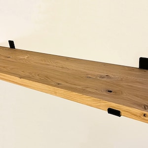 May include: A natural wood floating shelf with a light brown finish. The shelf is supported by four black metal brackets. The wood grain is visible, adding texture. This shelf is perfect for displaying items or storage.
