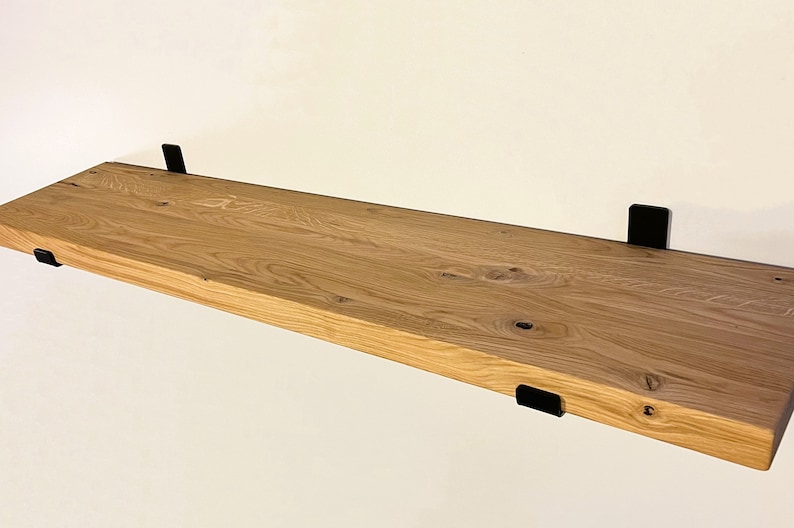 May include: A natural wood floating shelf with a light brown finish. The shelf is supported by four black metal brackets. The wood grain is visible, adding texture. This shelf is perfect for displaying items or storage.