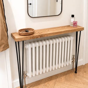 Peut inclure: Une console rectangulaire en bois au grain naturel, soutenue par quatre pieds fins en épingle à cheveux en métal noir. La table est placée au-dessus d'un radiateur à colonnes vertical blanc et porte un petit panier tressé avec des clés.