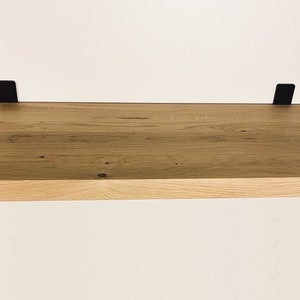 May include: A rectangular wooden shelf with a natural wood grain finish. The shelf is supported by four black metal brackets, two on the top and two on the bottom. The shelf is mounted on a white wall.