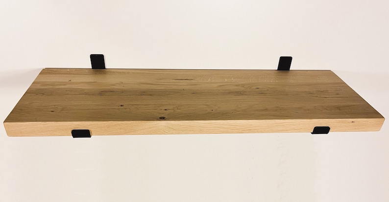 May include: A rectangular wooden shelf with a natural wood grain finish. The shelf is supported by four black metal brackets, two on the top and two on the bottom. The shelf is mounted on a white wall.