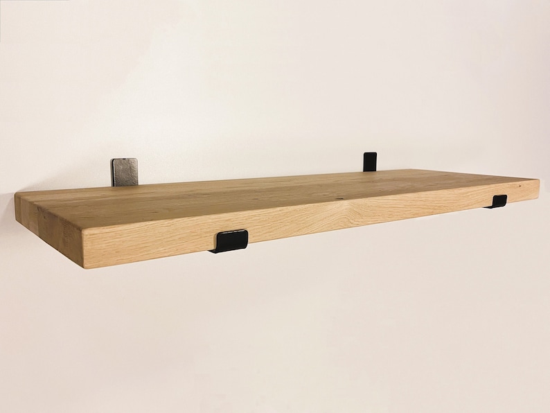 May include: A floating wooden shelf with a natural wood finish, supported by three black metal brackets. The shelf is rectangular and appears to be mounted on a white wall. The brackets are L-shaped and provide a modern, minimalist aesthetic.