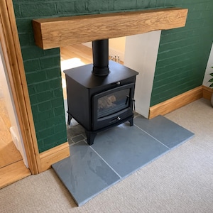 May include: A black metal wood-burning stove with a glass door sits on a gray stone hearth. Above the stove is a thick, natural-toned wooden mantel beam. The stove is positioned against a dark green painted brick wall with light-colored carpet.