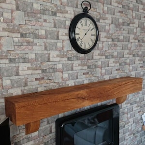May include: A wooden mantel shelf with a distressed finish, mounted above a black fireplace. The shelf is supported by two brackets. A black clock with Roman numerals is hanging on the wall above the shelf.