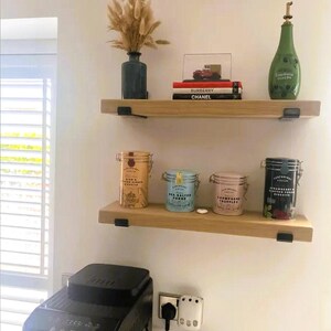 K&ouml;nnte beinhalten: Zwei Holzregale mit verschiedenen dekorativen Gegenst&auml;nden. Das obere Regal enth&auml;lt eine Vase mit getrockneten Blumen, B&uuml;cher und eine kleine Autofigur. Das untere Regal zeigt vier bunte Dosen. Die Regale sind an einer wei&szlig;en Wand befestigt.