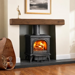 May include: A black cast iron wood-burning stove with a glass door showing bright orange flames, positioned under a thick dark brown wooden mantelpiece. Beside the stove sits a curved wooden log holder filled with several pieces of firewood.
