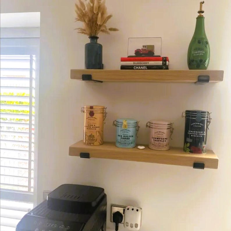 May include: Two wooden shelves with various decorative items. The top shelf holds a vase with dried flowers, books, and a green bottle. The bottom shelf displays four colorful tins. The shelves are mounted on a white wall.