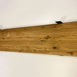 May include: A rectangular wooden shelf with a natural wood grain finish, mounted on a white wall. The shelf is supported by black metal brackets. The wood has visible knots and a warm, light brown color.