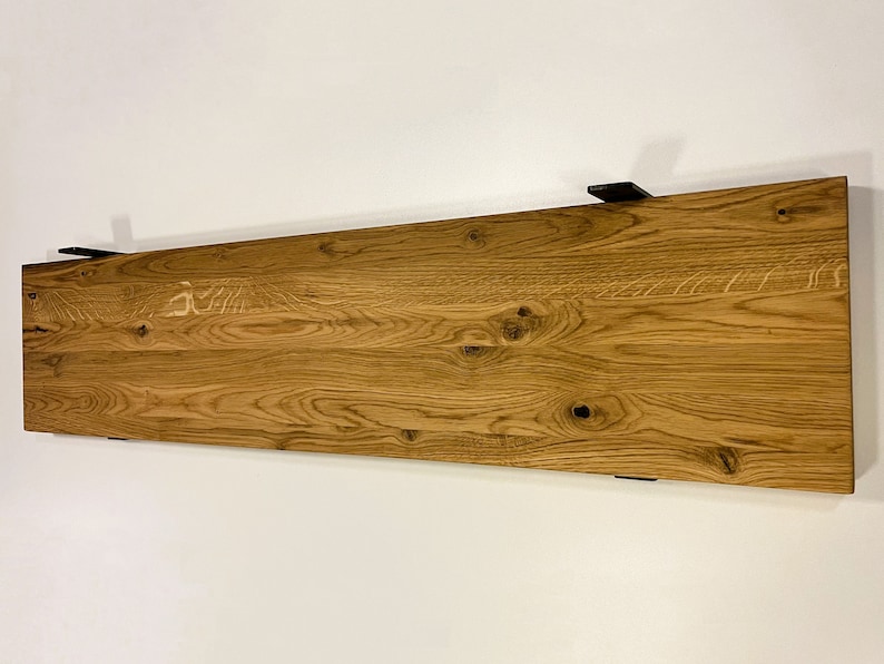 May include: A rectangular wooden shelf with a natural wood grain finish, mounted on a white wall. The shelf is supported by black metal brackets. The wood has visible knots and a warm, light brown color.