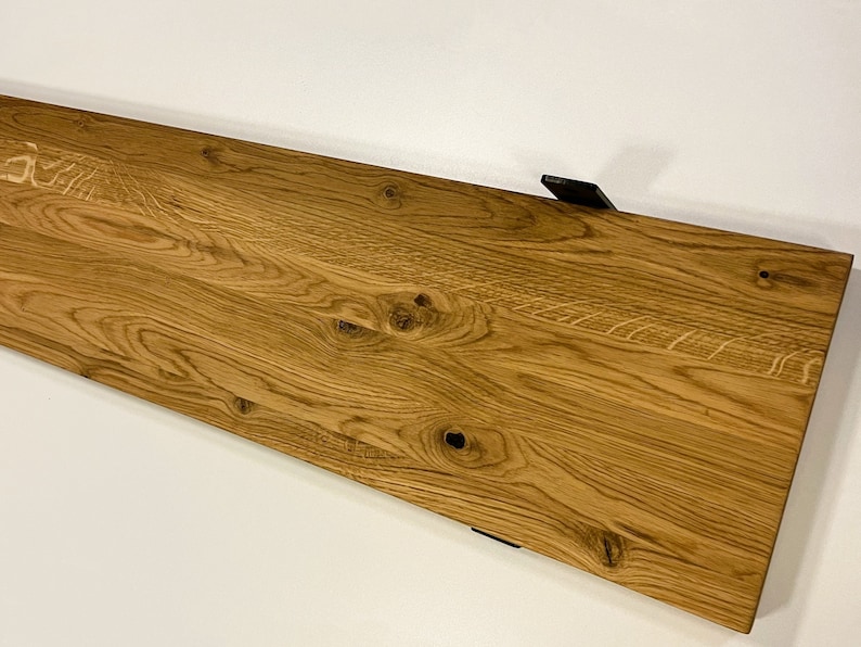 May include: A rectangular wooden shelf with a natural wood grain finish. The shelf is a warm, light brown color and features visible knots and grain patterns. Two black metal brackets are attached to the underside. The shelf is designed for wall mounting.