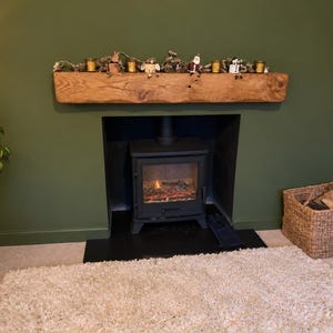 May include: A rustic wooden fireplace mantel mounted on a dark olive green wall above a black metal wood-burning stove. The mantel is decorated with a garland featuring small figurines, including a Santa Claus and various seasonal ornaments.