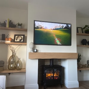 K&ouml;nnte beinhalten: Wohnzimmer mit Kamin, Fernseher und Einbauregalen. Der Kamin hat einen schwarzen Ofen mit Feuer. Der Fernseher zeigt eine Golfplatzszene. Regale mit B&uuml;chern, Dekoration und Vasen.