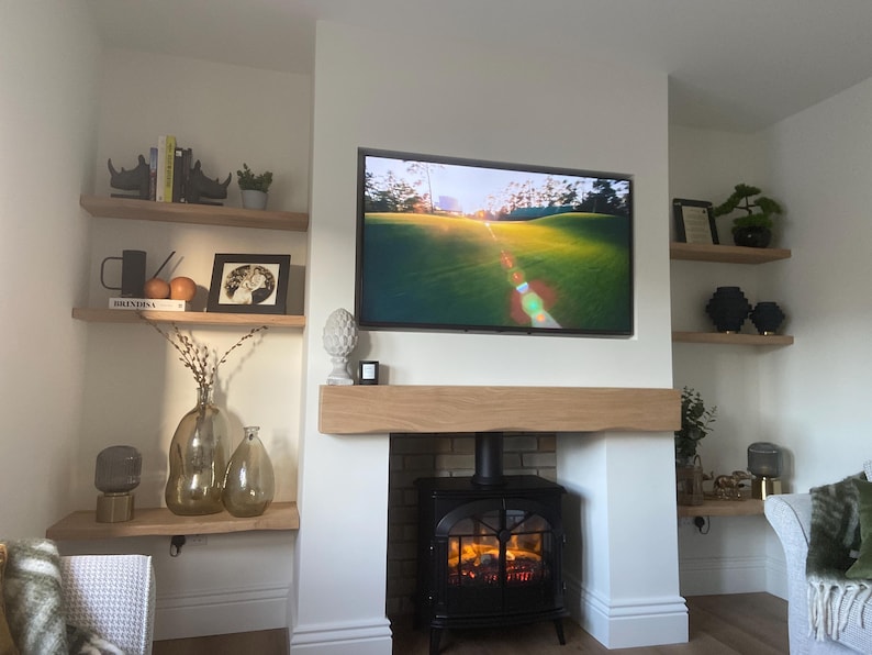 K&ouml;nnte beinhalten: Wohnzimmer mit Kamin, Fernseher und Einbauregalen. Der Kamin hat einen schwarzen Ofen mit Feuer. Der Fernseher zeigt eine Golfplatzszene. Regale mit B&uuml;chern, Dekoration und Vasen.