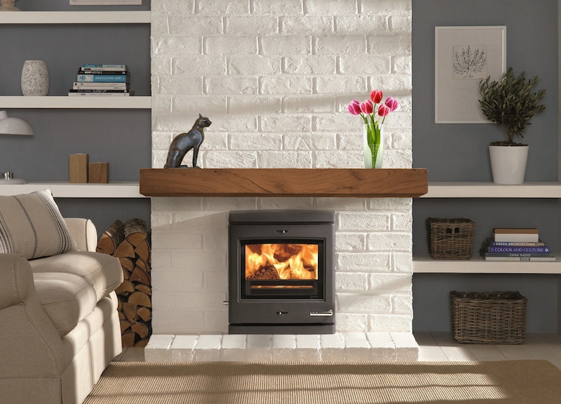 May include: A gray wood-burning stove with a glass door and a fire burning inside. The stove is set into a white brick fireplace with a wooden mantel. There is a black cat figurine on the mantel, and a vase of pink tulips.